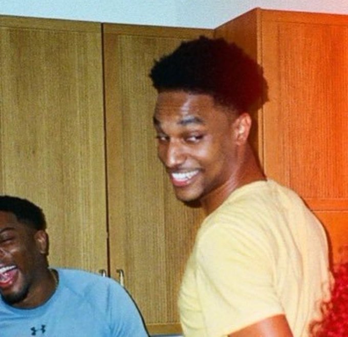A young man smiles broadly in a locker room while another person laughs next to him.
