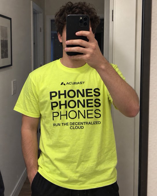 A person takes a mirror selfie wearing a neon yellow t-shirt that reads "ACURAST - PHONES PHONES PHONES - RUN THE DECENTRALIZED CLOUD."