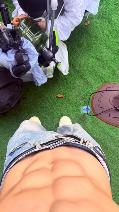 A shirtless man wearing jeans shows his abs in a mirror selfie while another person crouches nearby holding a camera on artificial turf. The image plays on the post's humor about missing a "male model event," with the photographer's gear suggesting a staged fashion-style shoot.