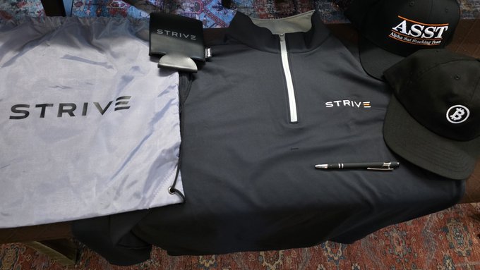 A spread of Strive-branded merchandise is displayed, including a white t-shirt with "STRIVE" printed across the chest, a black quarter-zip pullover with the Strive logo, a branded belt, a pen, and two hats—one featuring "ASST" (Asset Entities) and another with the Strive "B" logo—likely sent as a gift to a supporter who holds $ASST as their largest portfolio holding.