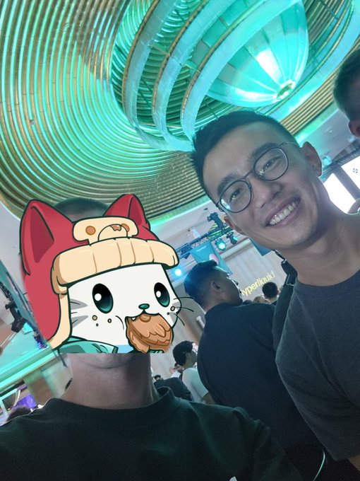 Two people pose for a selfie under an ornate teal-colored ceiling, with one person's face obscured by a cartoon cat filter.
