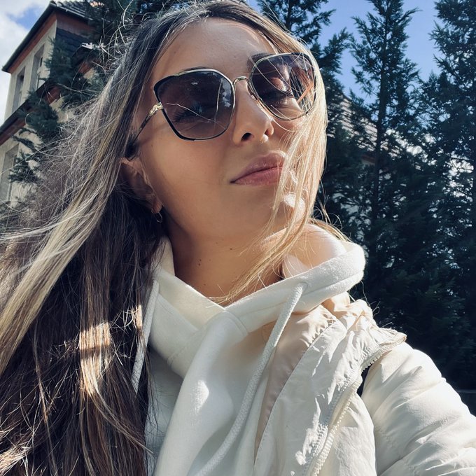A low-angle selfie of a woman with long brown hair wearing oversized sunglasses and a white puffy jacket, standing outdoors with pine trees and a building in the background.