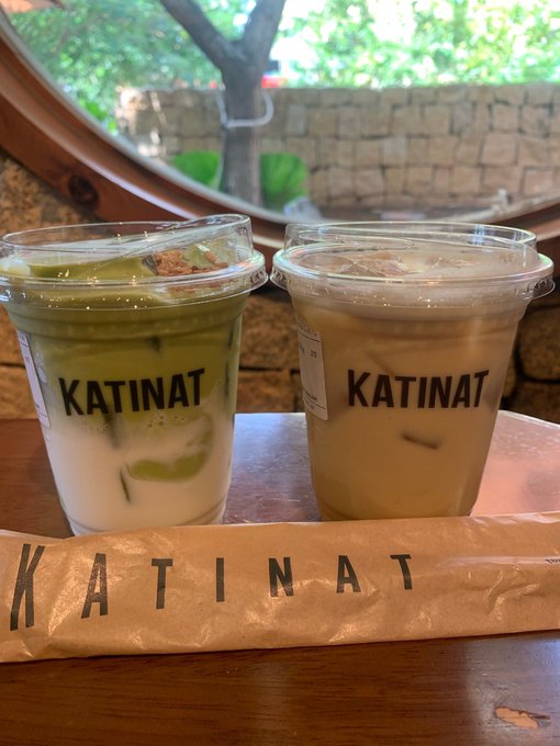 Two KATINAT iced drinks (a matcha latte and an iced coffee) and a paper-wrapped straw sit on a wooden table.