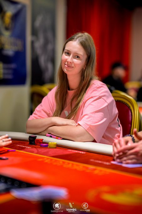 A smiling young woman with long blonde hair, wearing a pink t-shirt, sits at a red poker table with casino chips in front of her. The image features 'IMPERIUM', 'VAMOS POKER TOUR', and 'SERIOUS POKER' logos at the bottom.