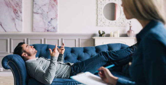 The image shows a man lying down on a blue couch, holding a pen and paper while looking at his reflection in a mirror. A woman is standing behind him, observing or possibly taking notes about the man's behavior. The scene suggests that they might be having a conversation or discussing something related to the man's actions or thoughts.