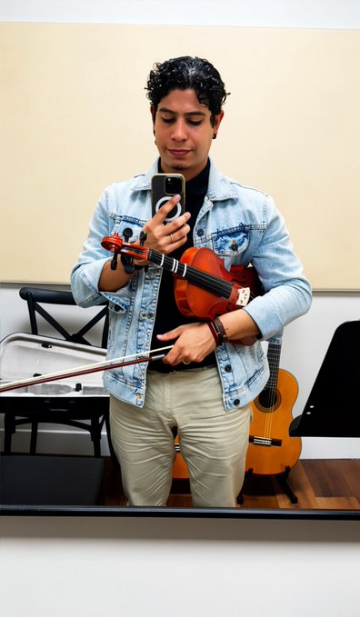 A mirror selfie of a young man wearing a denim jacket holding a violin and bow while taking the photo with his smartphone, with a guitar visible in the background.