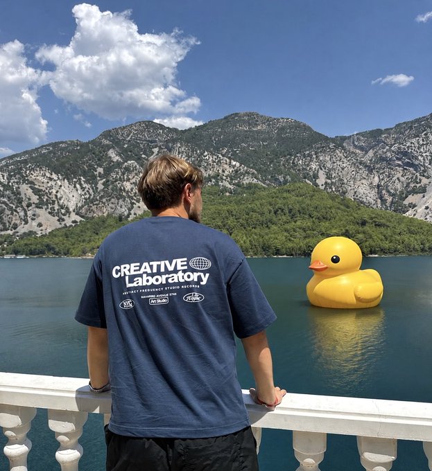 A person wearing a navy "Creative Laboratory" t-shirt leans on a white railing, looking out over a blue lake toward mountains while a giant yellow rubber duck floats on the water.
