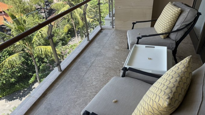 A balcony view showing outdoor seating with cushions, a white side table, and a tiled floor overlooking tropical palm trees.