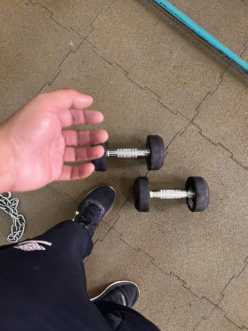 A first-person view looking down at a pair of black dumbbells resting on a gym floor, with the person's hand, black sneakers, and a chain visible.