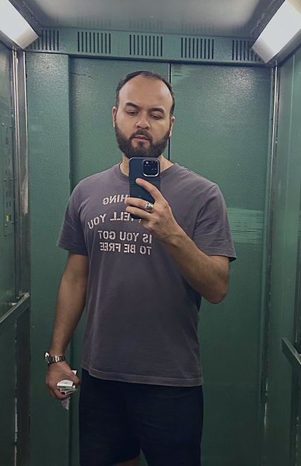 A bearded man takes a mirror selfie in a green-walled bathroom wearing a gray "TO BE FREE IS YOU GOT TO LET YOU THINK" graphic t-shirt, holding a phone and a small can. The casual mirror selfie visually reinforces the daily commitment to posting a selfie each day as part of the "$chog" price-tracking challenge.