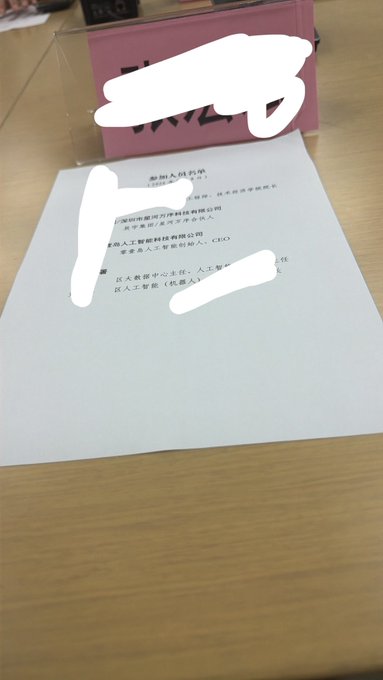 A handwritten note in Chinese on white paper placed on a wooden surface, with portions of the text and surrounding areas obscured by white digital redactions.