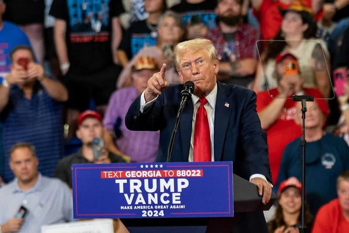 Former President Donald Trump speaking at a campaign rally behind a podium labeled 'TRUMP VANCE MAKE AMERICA GREAT AGAIN! 2024', with a sign reading 'TEXT GEORGIA TO 88022' and a crowd visible in the background.