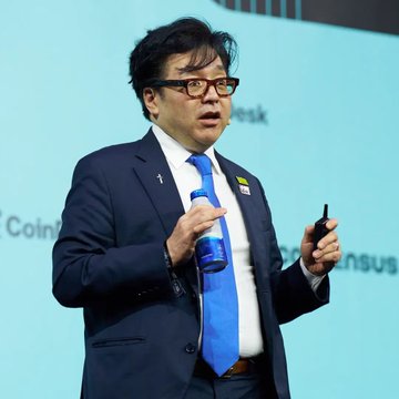 A man in a blue suit and tie speaks on stage while holding a water bottle and a presentation remote, with a "Coin" logo visible in the background.