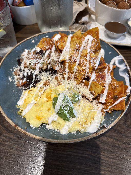 A close-up photo of a breakfast plate featuring scrambled eggs with peppers, tortilla chips covered in salsa and crema, and black beans, served on a blue ceramic dish with a glass of water nearby.