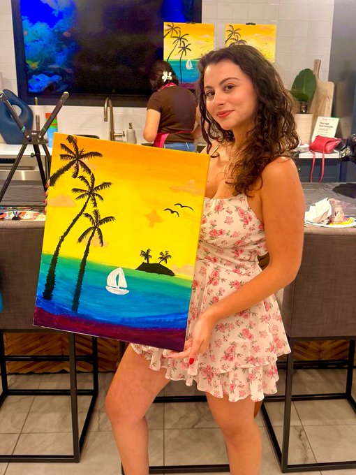 A woman in a floral dress proudly displaying a tropical sunset painting featuring palm trees and a sailboat at what appears to be a paint-and-sip class.