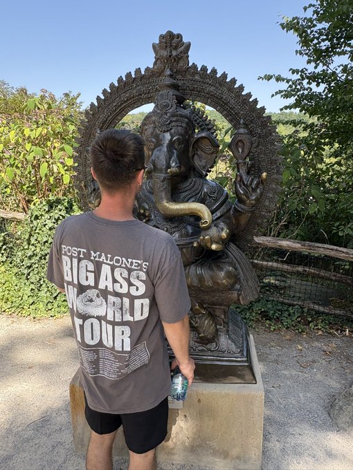 A young man wearing a 'POST MALONE'S BIG ASS WORLD TOUR' t-shirt stands facing a large bronze Ganesha statue outdoors.