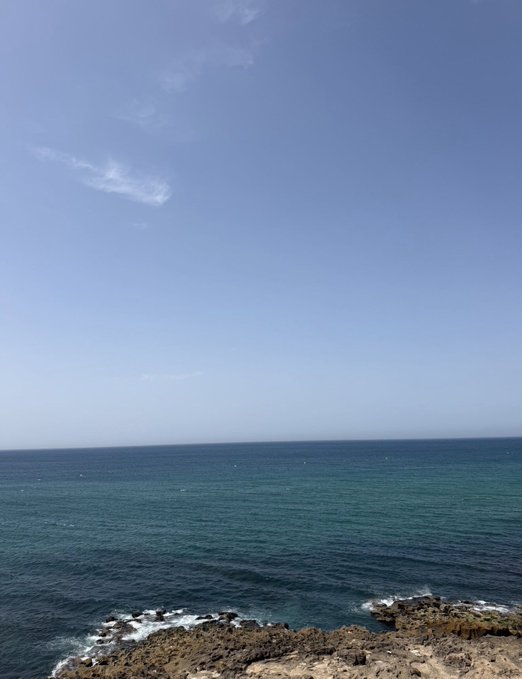 A scenic view of the ocean meeting a clear blue sky with wispy clouds, with rocky shoreline visible at the bottom of the frame.