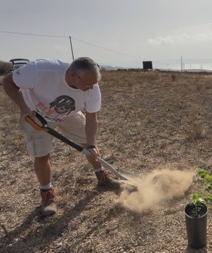 An older man wearing a white t-shirt, shorts, and a cap, using a pickaxe or shovel to dig a hole in dry, dusty ground, with a small potted plant visible in the foreground.