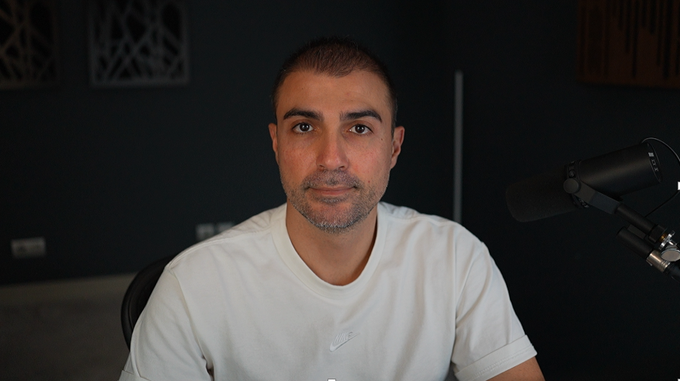 A man in a white Nike t-shirt sits in front of a microphone, announcing that a live YouTube broadcast has just started at youtube.com/live/4qXaRPOcl.