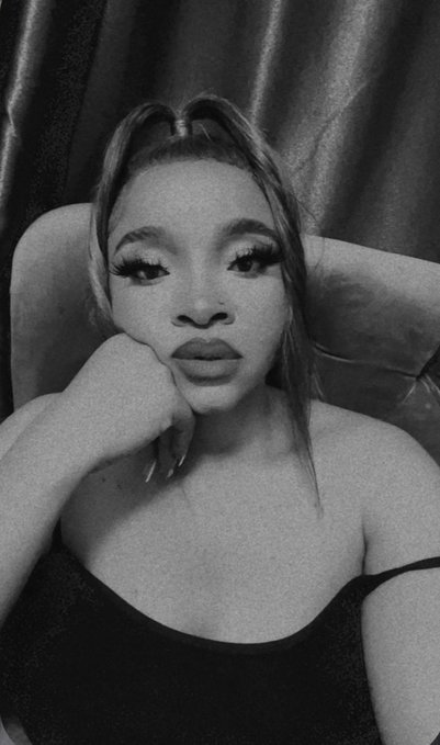 A black-and-white close-up portrait of a woman with dramatic eyelashes and full lips, resting her chin on her hand against a dark curtain backdrop.