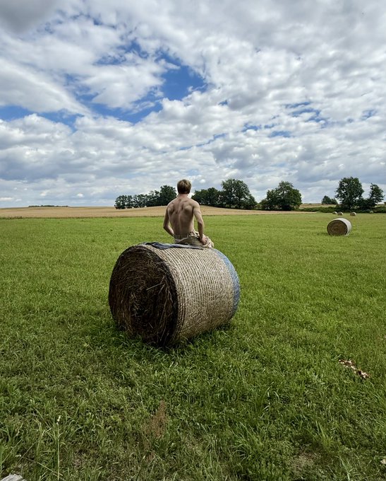 A shirtless man sits confidently atop a large round hay bale in an expansive green field, with another bale visible in the distance under a partly cloudy sky—mirroring the bold self-assuredness of the caption "I'm the best person I know." His relaxed yet commanding pose in the open landscape reflects a sense of quiet self-celebration.