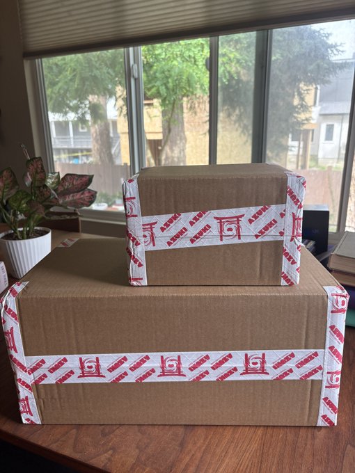 Two brown cardboard boxes sealed with red and white "Azuki" branded tape sit stacked on a wooden table, with the smaller box on top of the larger one. The image accompanies a post about the excitement of receiving an Azuki TCG order, showcasing the unopened delivery featuring the recognizable Azuki logo prominently displayed across the packing tape.