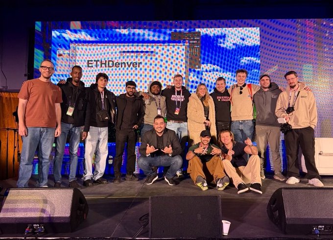 A group photo of attendees posing on stage at an 'ETHDenver' event with a colorful LED screen display behind them.