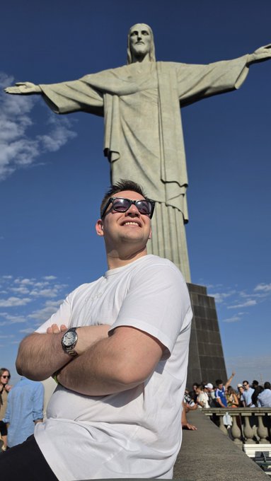 A smiling man in a white t-shirt and sunglasses poses in front of the iconic Christ the Redeemer statue in Rio de Janeiro on a sunny day.