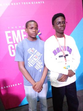 Two boys pose in front of a colorful "EMF" (Emerging Markets Forum or similar) branded backdrop featuring text like "WHERE NEXT TECH GIANTS" and "GANIZED BY," likely from a 2024 event where the older boy met the speaker and learned about "pxxl," now becoming part of his core team.