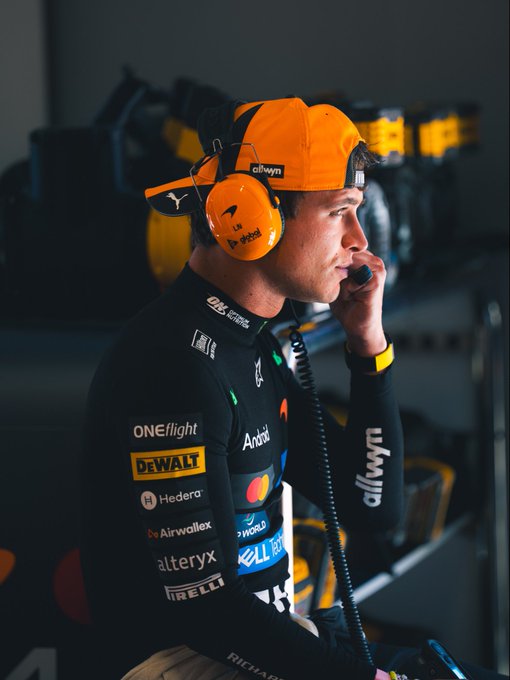 A focused McLaren F1 driver, wearing a black racing suit adorned with sponsor logos including DEWALT, Alteryx, Hilton, and Pirelli, holds a radio handset while sporting bright orange headphones and a matching cap emblazoned with "OKX" and "alwyn." The image captures a behind-the-scenes moment of preparation, perfectly aligning with the "Taking it all in" caption for the #SpanishGP.
