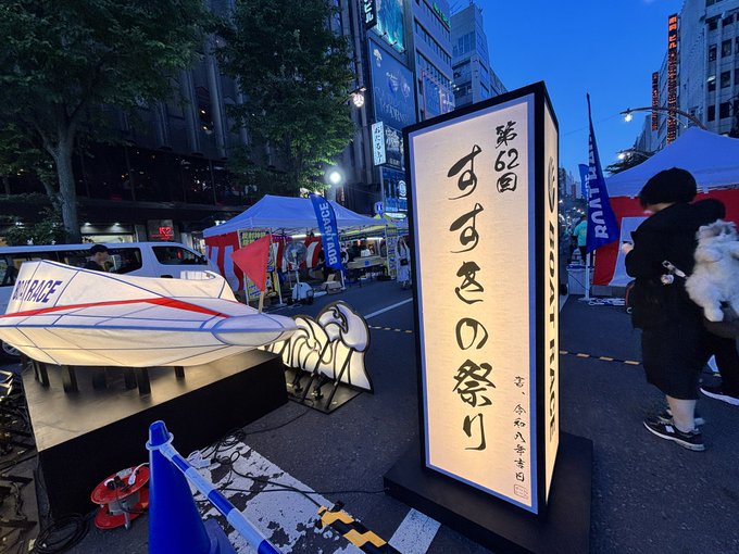 An illuminated Japanese sign reading '第62回 すすきの祭り' (62nd Susukino Festival) stands on a city street decorated with lanterns and festival stalls at dusk.