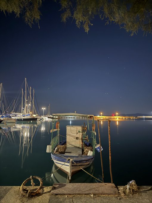 A fishing boat sits quietly moored at a harbor under a starry night sky, with distant lights reflecting across the calm water. The serene maritime scene pairs with the caption's playful hint that the boat was "bought with my moonpay airdrop," suggesting a whimsical claim of crypto-funded seaside purchases.