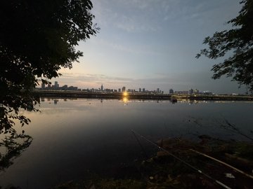 A tranquil sunrise over Chengdu's skyline reflects on the calm river waters, viewed from a quiet shoreline framed by silhouetted tree branches. The serene scene captures a peaceful morning fishing trip, contrasting with the post's playful note about "shrimp fishing" and enjoying the day despite recent losses.
