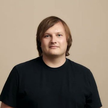 A portrait of a young man with light brown hair and bangs wearing a plain black t-shirt against a solid beige background.