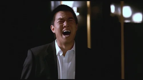 A man in a black suit and white shirt crying or shouting emotionally in a dimly lit setting with blurred lights behind him.