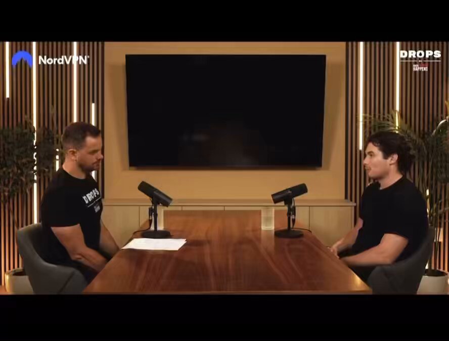 The image shows two men sitting at a table for a podcast recording session. They are both wearing black t-shirts and appear to be engaged in conversation, possibly discussing the performance of Gold over the past year or planning their strategy for the upcoming year. The background features a large flat screen TV mounted on the wall behind them, which could be used as an additional visual aid during their podcast discussions.