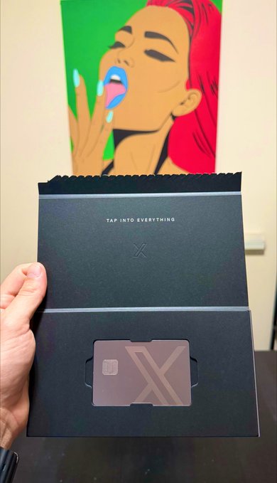 A hand holding an open black envelope with 'TAP INTO EVERYTHING' and 'X' printed on it, containing a card with an 'X' design, with a colorful pop-art portrait hanging on the wall behind it.