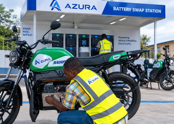 A worker in a yellow safety vest services a green and black 'Azura Swift' electric motorcycle at an Azura Battery Swap Station, with the station's motto 'Powering Africa's Mobility. Electrically.' and 'SWAP. RIDE. REPEAT.' visible.