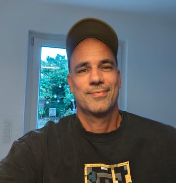 A selfie of a smiling bald man wearing a baseball cap and dark t-shirt, taken indoors with a window showing trees in the background.