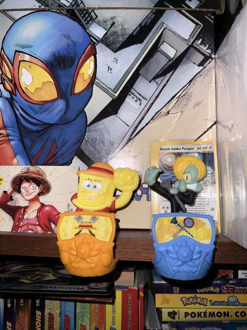Two SpongeBob SquarePants figurines (one orange/yellow with a straw hat, one blue with Squidward on top) displayed on a shelf with a Pokémon trading card and comic/manga books below.