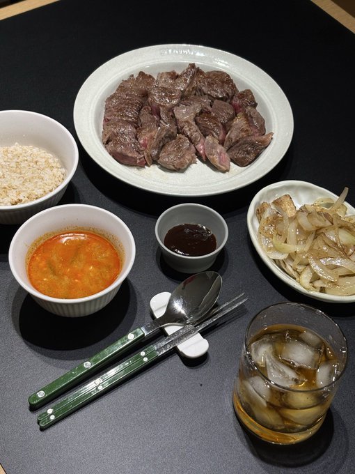 A hearty meal featuring grilled steak, rice, soup, sautéed onions, dipping sauce, and a glass of iced tea, paired with green-handled utensils on a dark placemat. The image reflects the post's message about eating well and staying healthy while patiently waiting for the crypto bull run to arrive.
