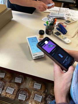 A person holding a smartphone displaying an app interface at what appears to be a market or shop counter, with jars of goods visible below.