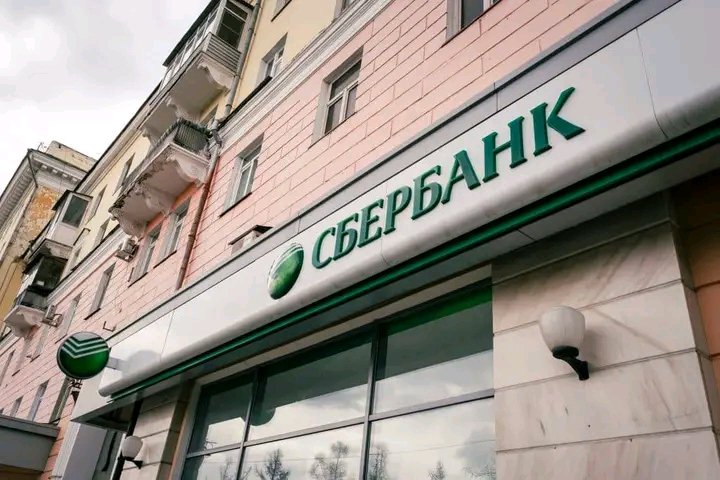 The exterior of a Sberbank (СБЕРБАНК) branch is shown, featuring the bank's green logo and Cyrillic signage on a modern storefront attached to a pink residential building.