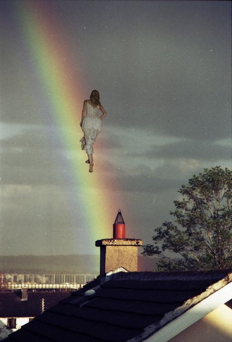 A surreal composite image showing a woman in a white dress floating in a starry night sky next to a rainbow, positioned above a rooftop chimney.