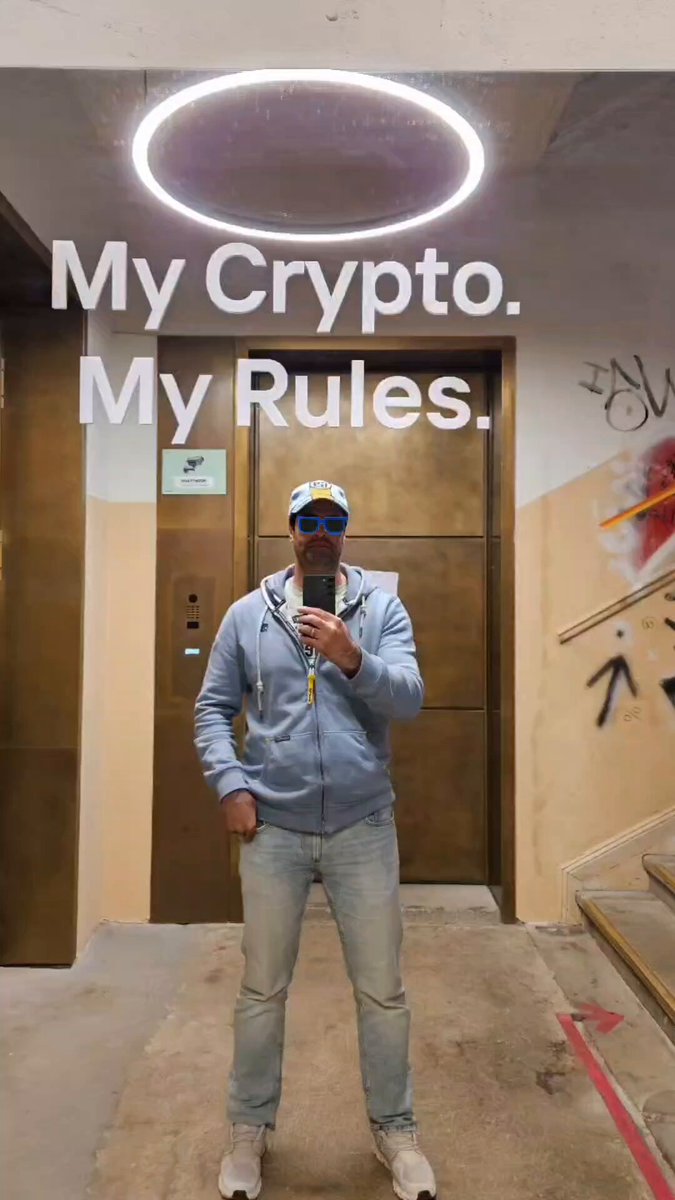 The image features a man standing in front of a door with "My Crypt. My Rules." written above him. The man appears to be taking a selfie using his cell phone, capturing this moment as part of his Blockchain Week experience.