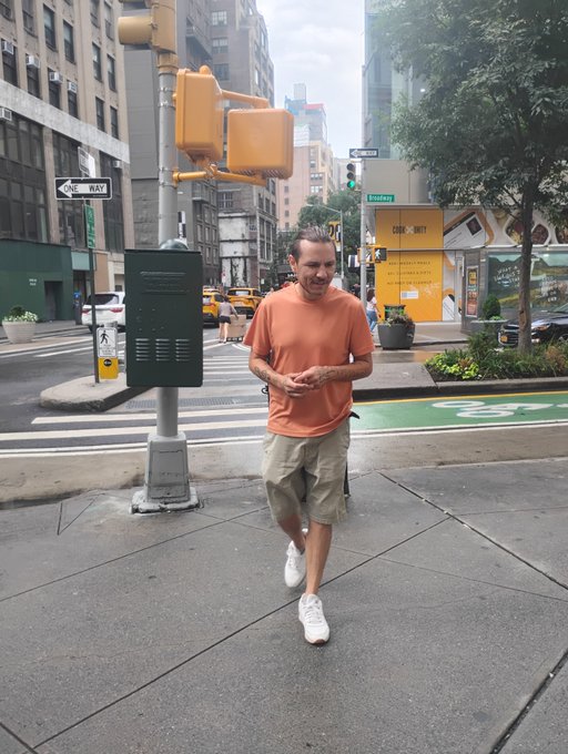 A candid photo captures a smiling man with his hair tied back, wearing an orange t-shirt and khaki shorts, walking on a city sidewalk near a crosswalk, a traffic light pole, and yellow taxis in the background.