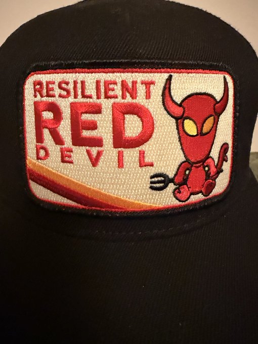 A black baseball cap displays a rectangular patch reading 'RESILIENT RED DEVIL' alongside an illustration of a small, cartoonish red devil character holding a pitchfork.