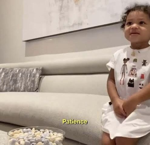 A young child wearing a white outfit with character prints sits calmly on a couch, with the word "Patience" displayed on screen. The caption humorously connects to the post about waiting until the last minute to request help, with "Patience" (likely meant to say "La Peace") playing on the child's composed demeanor contrasting with the community dev's likely panic.