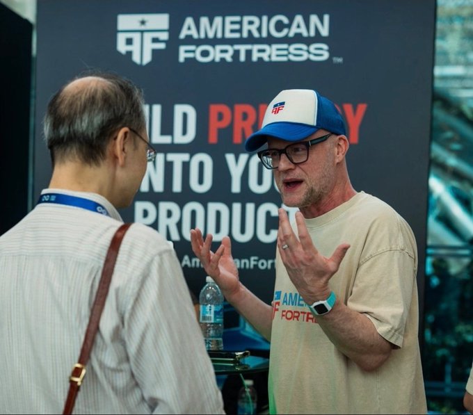 Two men conversing at an 'American Fortress' booth, with one wearing a branded cap and shirt, and a partially visible banner reading 'BUILD PROSPERITY INTO YOUR PRODUCT'.