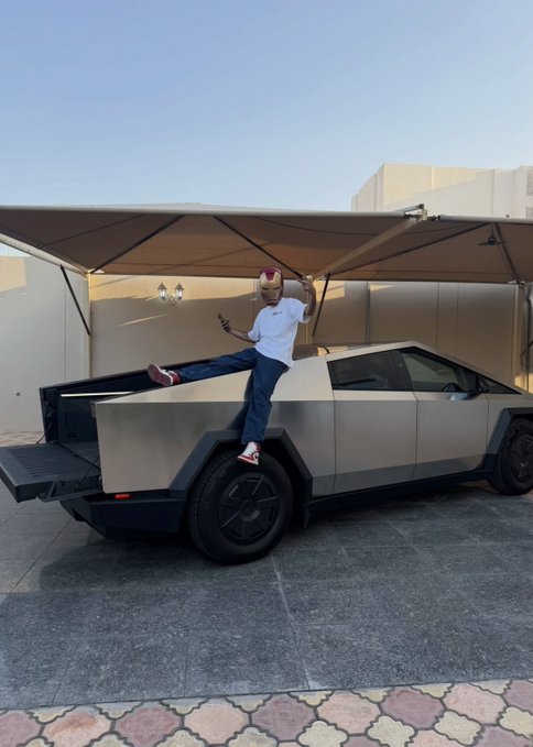 A man wearing pink bunny ears poses with one leg lifted against the bed of a silver Tesla Cybertruck parked under a carport.